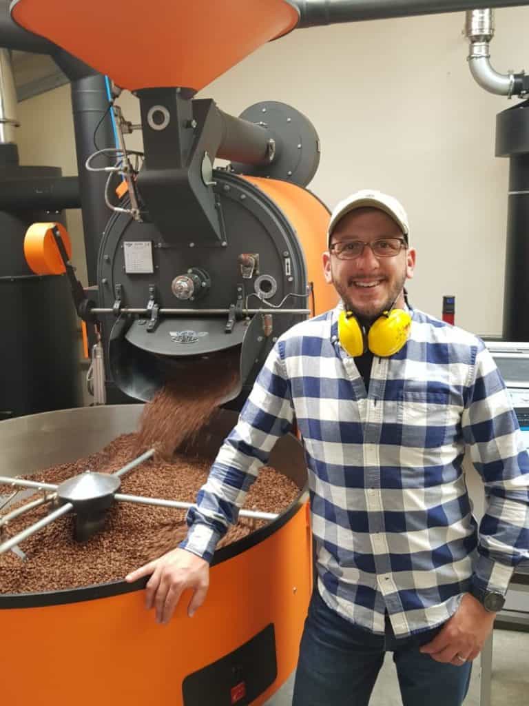 Badger and Dodo Coffee Roasters: Smiling Jacob, wearing a checked shirt and yellow headphones, stands beside an industrial coffee roaster as it releases freshly roasted beans into a large metal container. The modern facility buzzes with activity, welcoming each team member into the heart of the coffee-making process.