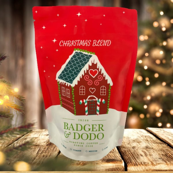 A festive red and white coffee bag labeled “Christmas Blend” from Badger & Dodo, featuring an illustration of a gingerbread house with candy cane pillars and a snowy roof. The bag sits on a wooden surface surrounded by pine branches and pinecones, creating a holiday atmosphere.