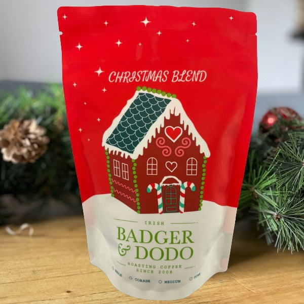 A festive red and white coffee bag labeled “Christmas Blend” from Badger & Dodo, featuring an illustration of a gingerbread house with candy cane pillars and a snowy roof. The bag sits on a wooden surface surrounded by pine branches and pinecones, creating a holiday atmosphere. A festive red and white coffee bag labeled “Christmas Blend” from Badger & Dodo, featuring an illustration of a gingerbread house with candy cane pillars and a snowy roof. The bag sits on a wooden surface surrounded by pine branches and pinecones, creating a holiday atmosphere.