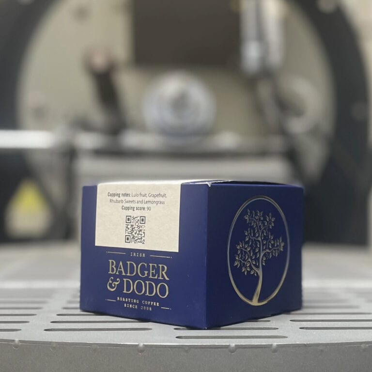 Blog 2 Badger & Dodo. A blue COLOMBIA–Jairo Arcila, Anaerobic coffee box from Badger & Dodo rests on a metal surface, evoking rich Colombian flavor, with a blurred machine in the background.