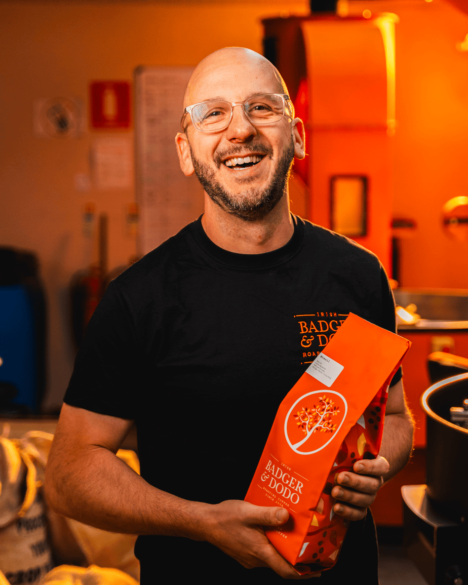 Badger and Dodo Coffee Roasters: A smiling person with glasses holds a bright orange bag of Badger & Dodo coffee in a warmly lit room, showcasing the quality trusted by wholesale partners, with coffee equipment and bags visible in the background.