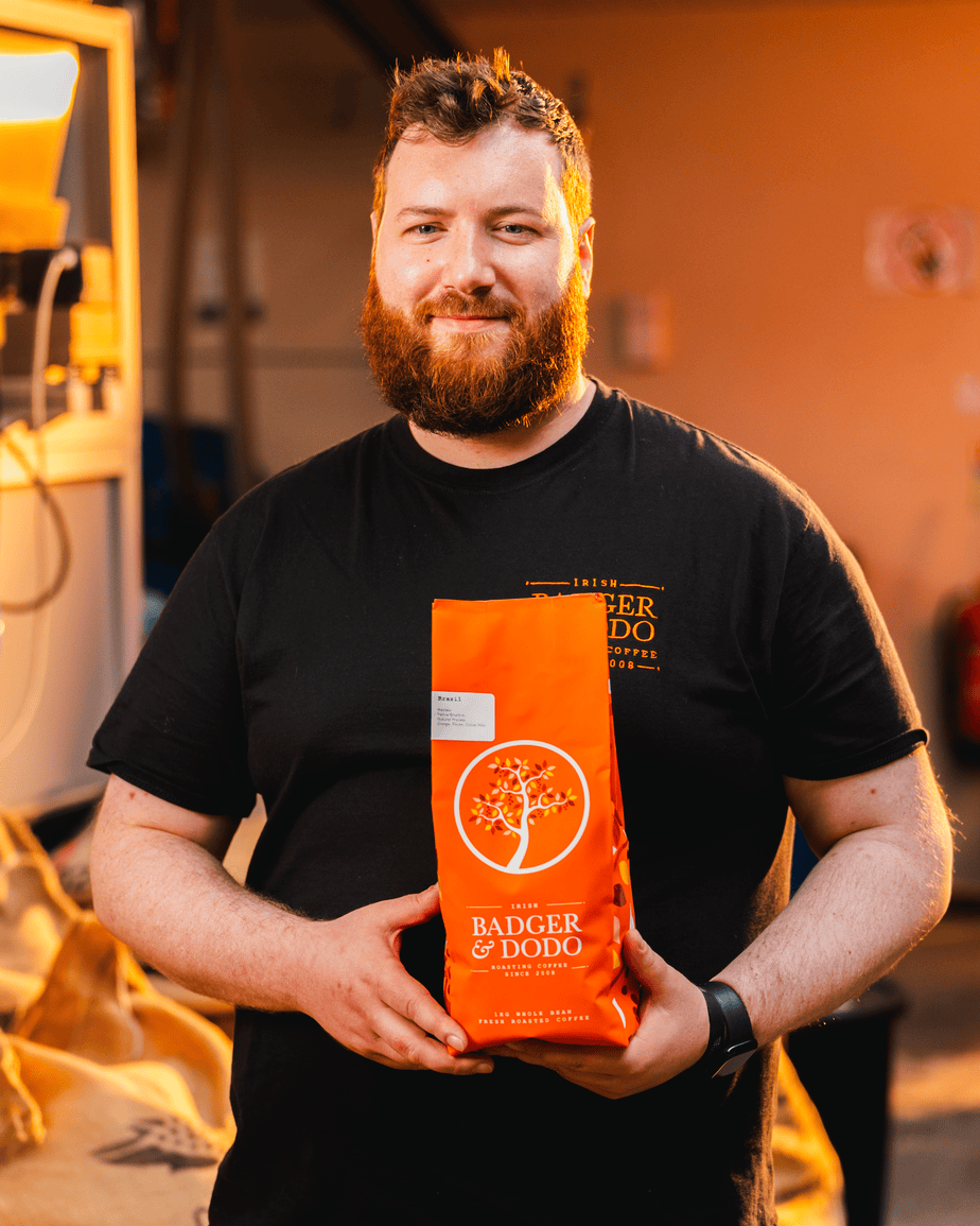 Badger and Dodo Coffee Roasters: A person with a beard smiles while holding a bright orange coffee bag labeled Badger & Dodo. They stand indoors, wearing a black T-shirt with the same logo, likely as a wholesale partner. The background appears to be a bustling coffee roasting facility.