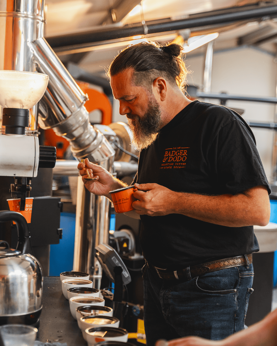 Badger and Dodo Coffee Roasters: A man with a beard and a bun, wearing a black shirt, is focused on tasting coffee from cups arranged on a table. In the industrial settings metal piping backdrop, he evaluates each blend with precision—an essential task for their wholesale partner selection.