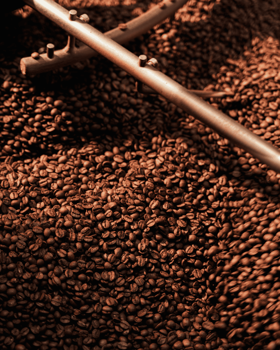 Badger and Dodo Coffee Roasters: Close-up of a large batch of roasted coffee beans being stirred by a metal arm in a commercial roasting machine. The lighting creates a warm, rich tone, highlighting the texture and color—perfect for enticing any wholesale partner seeking premium quality.