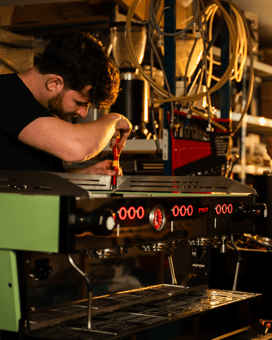 Badger and Dodo Coffee Roasters: In a dimly lit workshop, a wholesale partner skillfully uses a screwdriver to adjust a large green espresso machine with red digital displays and a metallic finish. Shelves brimming with equipment fill the background.