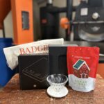 A coffee brewing set displayed on a wooden surface, featuring a black hand coffee grinder, and a red bag of Badger & Dodo Christmas Blend coffee with a gingerbread house design. Industrial coffee roasting equipment is visible in the blurred background.