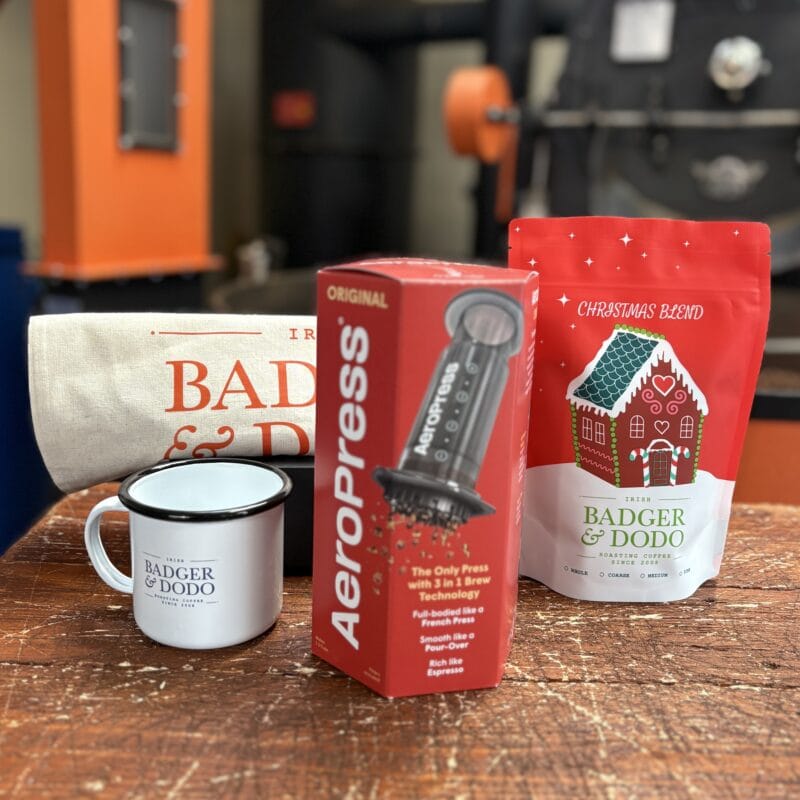 Badger & Dodo. The On The Go Bundle is a coffee gift set featuring Badger & Dodo products, an AeroPress Starter kit, mug, tote bag, and a red bag of Badger & Dodo Christmas Blend coffee with a gingerbread house design. Industrial coffee roasting equipment is visible in the blurred background—ideal for coffee lovers with busy lifestyles.