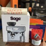 Badger & Dodo. The Sage Bundle is a coffee gift set featuring Badger & Dodo products, a Sage Barista Express, tote bag, a red bag of Badger & Dodo Christmas Blend coffee with a gingerbread house design. Industrial coffee roasting equipment is visible in the blurred background—ideal for coffee lovers with busy lifestyles.