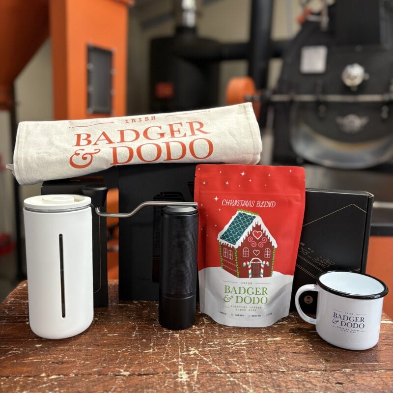 A coffee-themed gift set displayed on a wooden surface. The items include a white towel labeled “Badger & Dodo,” a red bag of “Badger & Dodo Christmas Blend” coffee with a gingerbread house design, a white and black manual coffee grinder, a white coffee mug with the “Badger & Dodo” logo, and other coffee accessories. The background shows coffee roasting equipment in an orange and black setting.