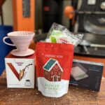 A coffee brewing setup displayed on a wooden surface, featuring a white a pink V60 dripper, V60 Range server, V60 Filters, Scales and a red bag of Badger & Dodo Christmas Blend coffee with a gingerbread house design on the packaging. The background shows coffee roasting equipment in an industrial setting.