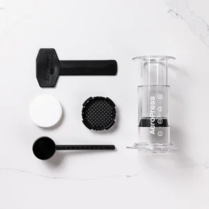 Badger and Dodo Coffee Roasters: A disassembled Aeropress Coffee Maker-Clear Colours is displayed on a white surface, featuring the main chamber with plunger, scoop, funnel, stirring paddle, and filter cap.