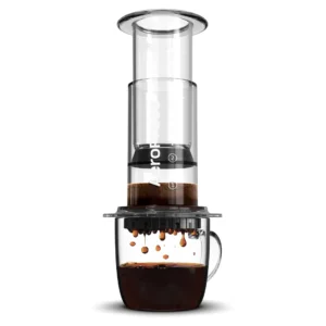 Badger and Dodo Coffee Roasters: A clear Aeropress Coffee Maker-Clear Colours is set over a glass mug, with coffee dripping through its filter. The transparent design highlights the vertically stacked components as it brews the perfect cup.