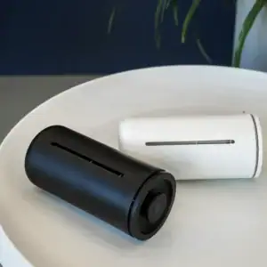 Two modern, cylindrical French press coffee makers in black and white, lying on their sides on a white round tray. Each has a vertical window showing the liquid level inside.