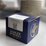 A navy blue box of Badger & Dodo coffee with gold detailing and logo, sitting on a white surface. A white label on top displays cupping notes: "Raspberry, Chocolate, Floral, Passion Fruit" and a cupping score of 89. A QR code is also visible on the label. The packaging has a premium, artisanal appearance.