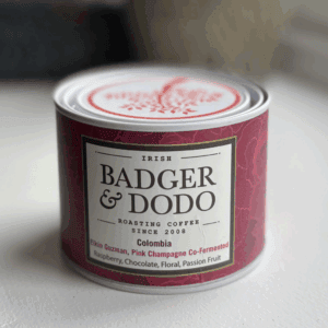 A cylindrical tin of Badger & Dodo coffee with a deep magenta label. The label reads "Irish Badger & Dodo Roasting Coffee Since 2008" and describes the contents as "Colombia – Elkin Guzman, Pink Champagne Co-Fermented" with flavor notes of raspberry, chocolate, floral, and passion fruit. The tin features a decorative lid with a red tree design.