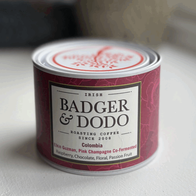 Blog 1 A cylindrical tin of Badger & Dodo coffee with a deep magenta label. The label reads "Irish Badger & Dodo Roasting Coffee Since 2008" and describes the contents as "Colombia – Elkin Guzman, Pink Champagne Co-Fermented" with flavor notes of raspberry, chocolate, floral, and passion fruit. The tin features a decorative lid with a red tree design.