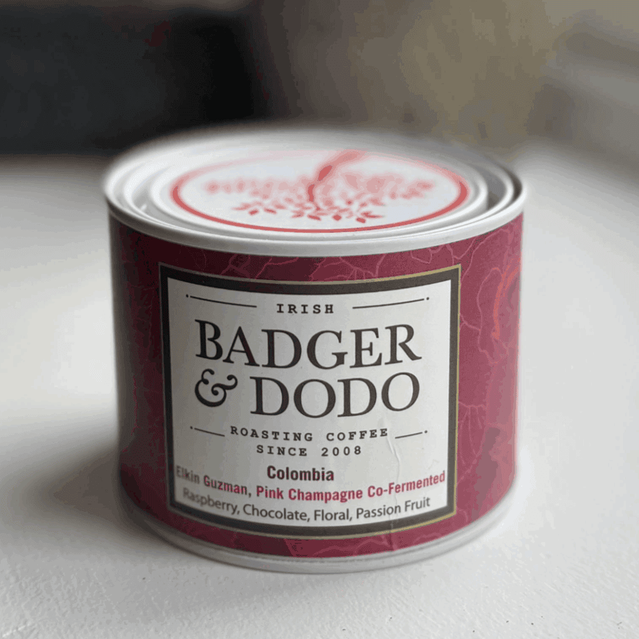 A cylindrical tin of Badger & Dodo coffee with a deep magenta label. The label reads "Irish Badger & Dodo Roasting Coffee Since 2008" and describes the contents as "Colombia – Elkin Guzman, Pink Champagne Co-Fermented" with flavor notes of raspberry, chocolate, floral, and passion fruit. The tin features a decorative lid with a red tree design.