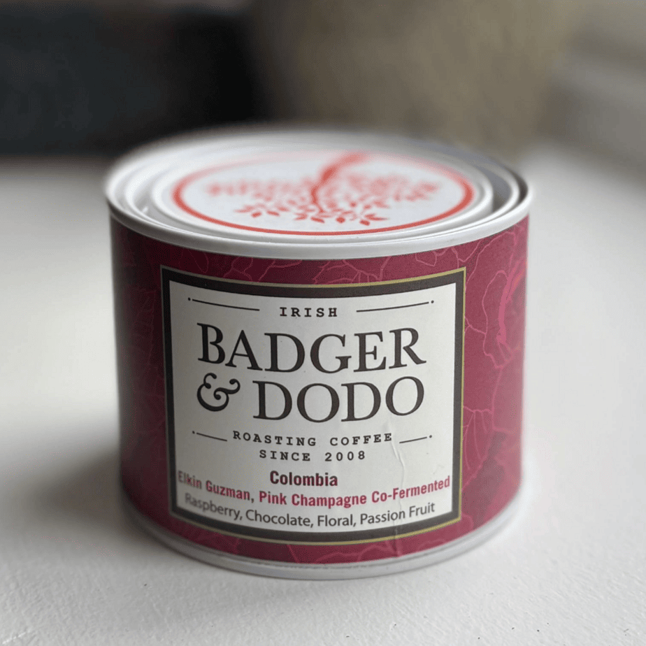 A cylindrical tin of Badger & Dodo coffee with a deep magenta label. The label reads "Irish Badger & Dodo Roasting Coffee Since 2008" and describes the contents as "Colombia – Elkin Guzman, Pink Champagne Co-Fermented" with flavor notes of raspberry, chocolate, floral, and passion fruit. The tin features a decorative lid with a red tree design.