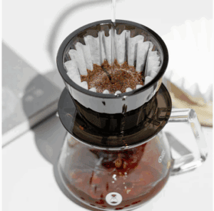 A top-down view of a pour-over coffee setup with hot water being poured over ground coffee in a paper filter inside a black plastic dripper, which is placed on top of a glass carafe partially filled with brewed coffee.