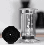 An AeroPress coffee maker is shown standing upright on a counter with a transparent chamber labeled with numbers 1 to 4. Next to it lies a black puck-shaped accessory with a textured honeycomb-like surface and a central hole, likely an attachment or filter cap. The background is softly blurred, showing a kitchen setting.