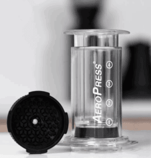 An AeroPress coffee maker is shown standing upright on a counter with a transparent chamber labeled with numbers 1 to 4. Next to it lies a black puck-shaped accessory with a textured honeycomb-like surface and a central hole, likely an attachment or filter cap. The background is softly blurred, showing a kitchen setting.