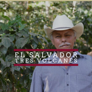 A smiling older man wearing a white cowboy hat and a checkered shirt stands in front of lush green coffee plants. Overlaid text reads “El Salvador Tres Volcanes” in bold white letters, framed by two horizontal maroon bars.