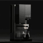 A modern black coffee machine dispensing coffee into a white dripper placed above a glass carafe. The machine is shown in a dark setting with soft lighting highlighting its sleek design and control knobs.
