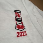 The image shows a close-up view of the embroidered design on a white T-shirt. The design features a red and black illustration of an AeroPress coffee maker, with a stylized coffee bed inside a hexagon at the bottom. Below the graphic, the year “2025” is stitched in bold red text.