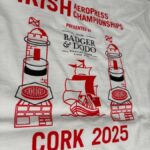 The image shows a close-up of the back design on a white T-shirt for the Irish Aeropress Championships. The top features the bold red text “IRISH Aeropress Championships” and the phrase “Presented by Badger & Dodo Roasting Coffee Since 2008” in black. Below, there are two stylized red-and-black AeroPress illustrations positioned like lighthouses, one on each side of a red sailing ship graphic. At the bottom, large red text reads “CORK 2025.”