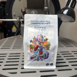 A white bag of Badger & Dodo coffee sits on the metal tray of a coffee roaster. The bag is labeled “2026 Irish AeroPress Championships” and features colorful artwork of people, tropical plants, and a dragon-like creature. A black roasting machine and lamp are visible in the background.