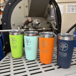 Four colorful 10oz YETI Rambler Tumblers displayed on a metal surface, each laser-etched with the Badger & Dodo logo and branding. The tumblers are in Green, mint green, bright orange, and navy blue, shown in a workshop setting with machinery and a lamp in the background.