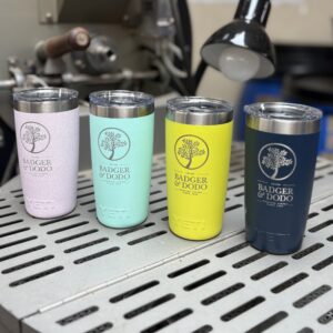 Four colorful 10oz YETI Rambler Tumblers displayed on a metal surface, each laser-etched with the Badger & Dodo logo and branding. The tumblers are in pastel pink, mint green, bright yellow, and navy blue, shown in a workshop setting with machinery and a lamp in the background.