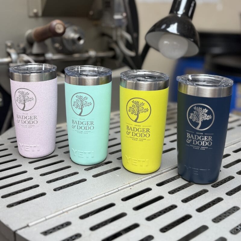 Four colorful 10oz YETI Rambler Tumblers displayed on a metal surface, each laser-etched with the Badger & Dodo logo and branding. The tumblers are in pastel pink, mint green, bright yellow, and navy blue, shown in a workshop setting with machinery and a lamp in the background.