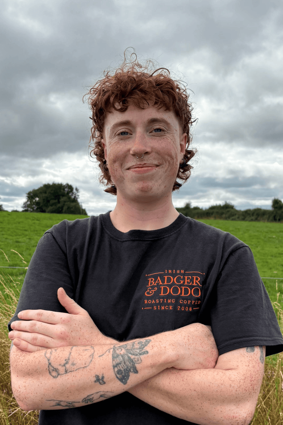 Person with curly red hair standing in a grassy field, wearing a black “Badger & Dodo” t-shirt and black shorts with a white drawstring. They are smiling with arms crossed, showing tattoos on their forearms. The sky is cloudy in the background.