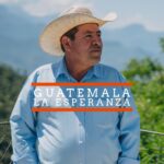 A man wearing a light blue button-up shirt and a straw hat stands outdoors, looking to the side. Behind him are blurred green trees and distant mountains under a clear sky. Text over the image reads "GUATEMALA LA ESPERANZA" with orange lines above and below "GUATEMALA."