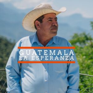 A man wearing a light blue button-up shirt and a straw hat stands outdoors, looking to the side. Behind him are blurred green trees and distant mountains under a clear sky. Text over the image reads "GUATEMALA LA ESPERANZA" with orange lines above and below "GUATEMALA."