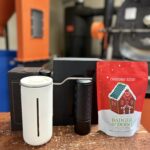 A coffee brewing setup displayed on a wooden surface, featuring a white French press, a black hand coffee grinder, and a red bag of Badger & Dodo Christmas Blend coffee with a gingerbread house design on the packaging. The background shows coffee roasting equipment in an industrial setting.