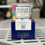 A small cylindrical tin of Badger & Dodo coffee sits atop a matching blue box. The tin features colorful illustrations of pineapples and tropical foliage, along with a label indicating it is a Colombian coffee from Felipe Arcila in Armenia, Quindio, with tasting notes of sugar cane, pineapple, papaya, and tea-like flavors. The box below displays the Badger & Dodo logo and includes a white label with cupping notes, a cupping score of 89, and a QR code. The items rest on a metal surface with a blurred background.