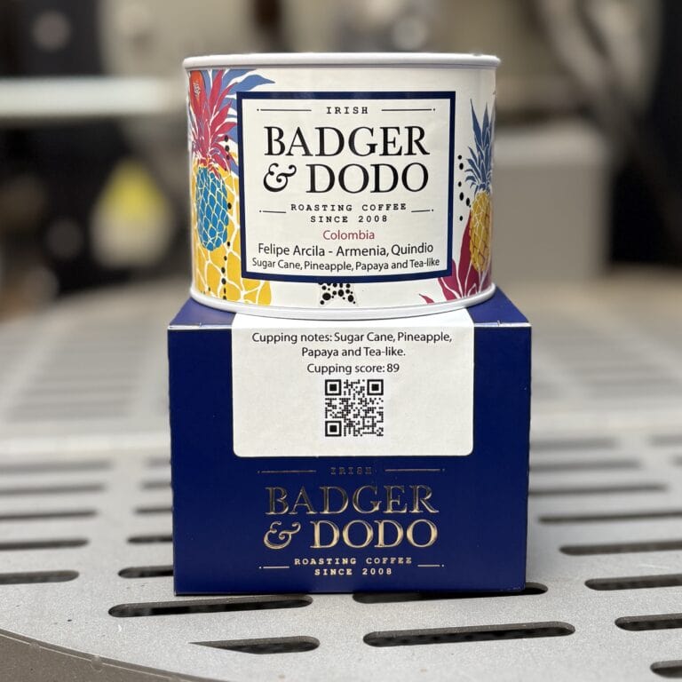 A canister of Badger & Dodo coffee sits on top of its matching retail box. The canister has a colorful design featuring illustrated pineapples and leaves, with a label that reads “Irish Badger & Dodo Roasting Coffee Since 2008 – Colombia, Felipe Arcila – Armenia, Quindio. Sugar Cane, Pineapple, Papaya and Tea-like.” The box below is dark blue with gold Badger & Dodo branding and a white label showing cupping notes and a QR code. The items are placed on a metal surface with a softly blurred background.