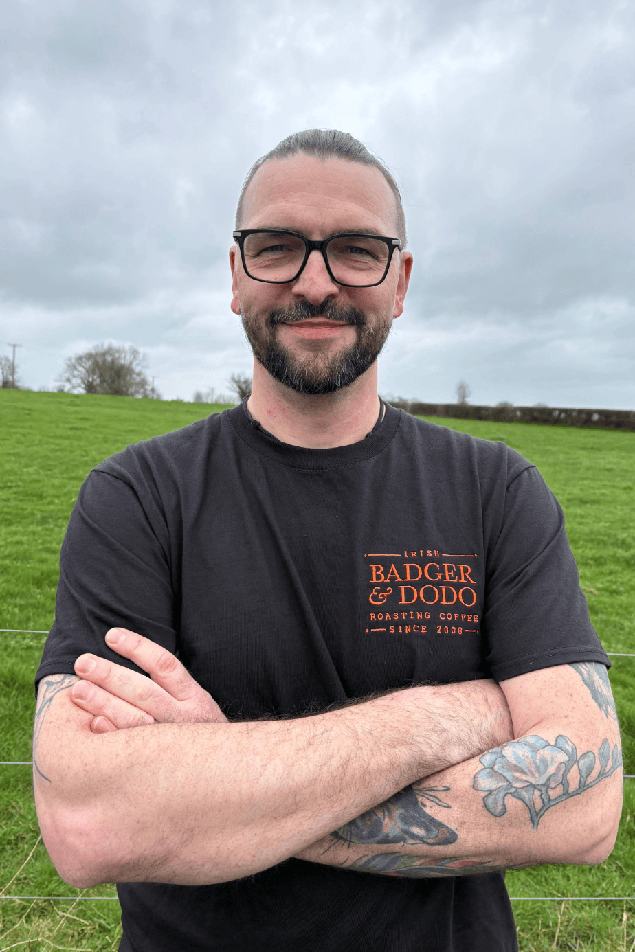 A smiling man wearing glasses and a black “Badger & Dodo” coffee T-shirt stands outdoors in a green field with his arms crossed, showing colorful forearm tattoos under a cloudy sky.