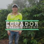A man wearing a bright yellow shirt and a blue cap stands in a lush coffee plantation surrounded by green plants and ripening coffee cherries. The setting appears misty or foggy, suggesting a highland environment. Text overlaid on the image reads “Ecuador – Marco Morocho.”