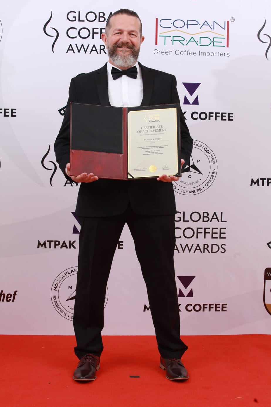 A man in a black tuxedo stands on a red carpet, smiling while holding an open certificate folder. Behind him is a branded backdrop for the Global Coffee Awards with various sponsor logos.