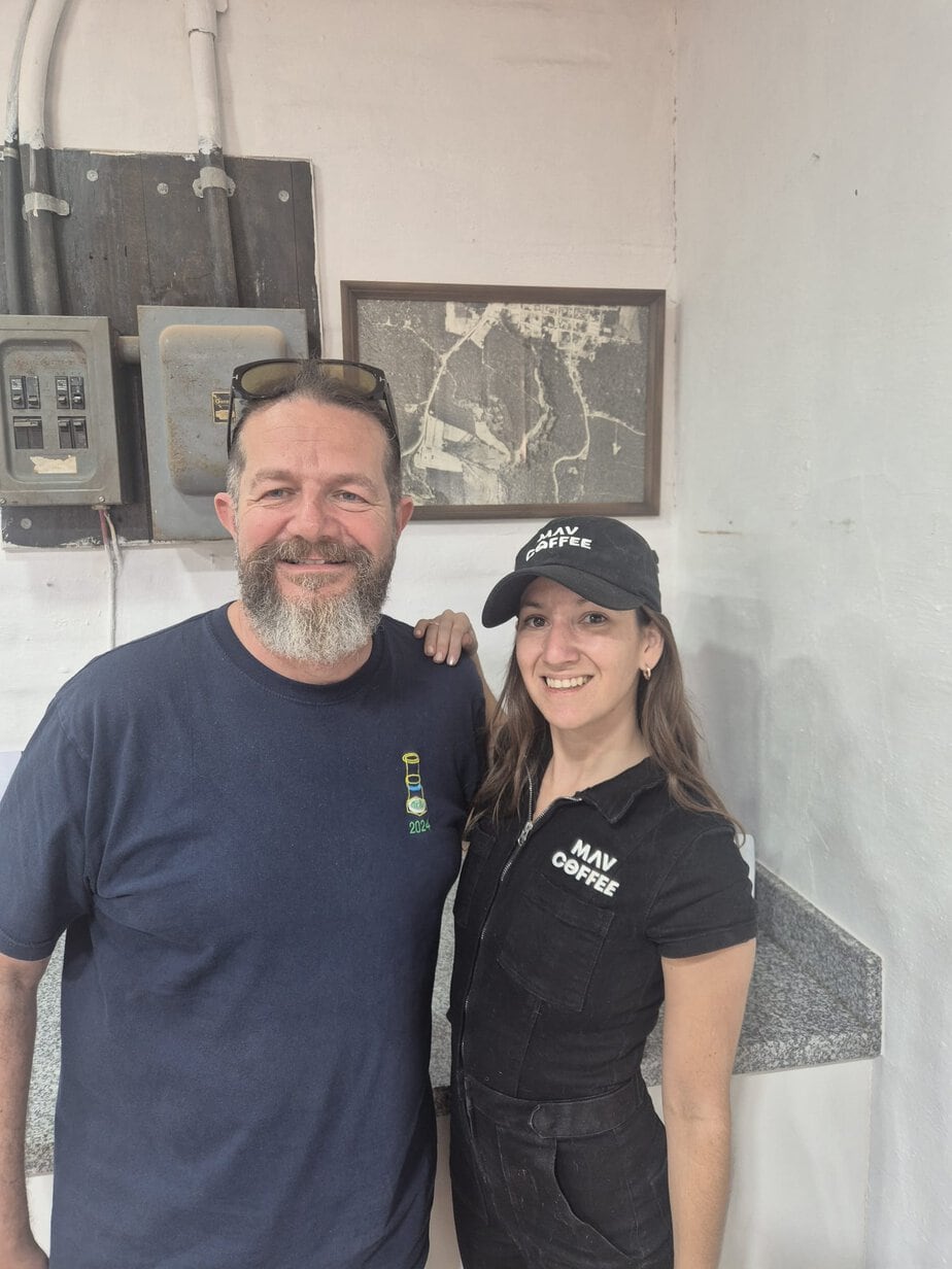 Two people smiling together inside a coffee shop or roastery, with a woman wearing MAV Coffee branded uniform and cap.