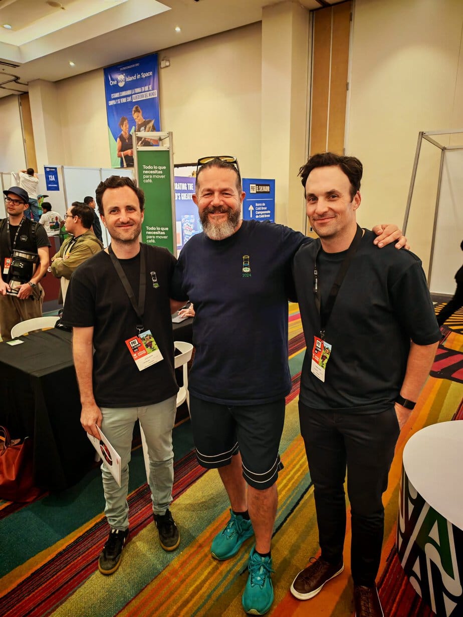 Three men posing together and smiling at what appears to be a coffee industry trade show or expo, with vendor booths and signage visible in the background.
