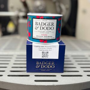 Alt text: A colorful tin of Badger & Dodo coffee with a cherry pattern sits on top of a blue branded box on a metal surface. The label reads “Irish Badger & Dodo Roasting Coffee Since 2008,” with details “Colombia Jairo Arcila – Santa Monica” and flavor notes including maraschino cherry, cola bottles, and amaretto. A sticker on the box shows cupping notes, a score of 88.5, and a QR code.