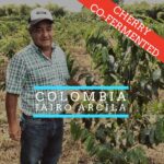 A man wearing a cap and a green plaid shirt stands in a coffee farm field surrounded by rows of coffee plants. Overlaid text reads “Colombia Jairo Arcila,” with a red banner in the corner that says “Cherry Co-Fermented.”