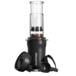 Black AeroPress coffee maker with a clear brewing chamber and plunger assembled on top of an insulated travel tumbler. A black lid and measuring scoop are placed beside the tumbler, all displayed against a white background.