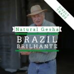 Promotional coffee banner featuring an older man wearing a straw hat and striped shirt, standing outdoors and smiling. Overlaid text reads “Natural Gesha” and “Brazil Brilhante,” with a diagonal banner in the corner stating “Direct Trade.” The design uses green and white typography against a darkened background.