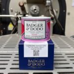 Badger & Dodo Colombia Jario Arcila coffee tin with pink watermelon-print label, sitting atop its navy blue box printed with cupping notes of Watermelon Candy, Key Lime, Banana, and Potpourri, in front of a coffee roaster