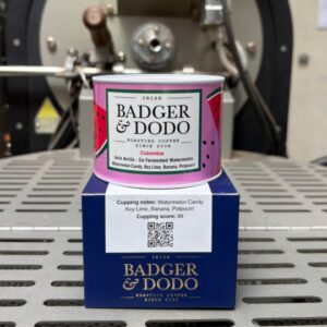 Badger & Dodo Colombia Jario Arcila coffee tin with pink watermelon-print label, sitting atop its navy blue box printed with cupping notes of Watermelon Candy, Key Lime, Banana, and Potpourri, in front of a coffee roaster